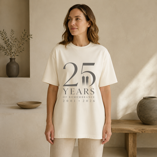A woman wearing an ivory-colored oversized t-shirt with a dark gray graphic that reads '25 YEARS OF REMEMBRANCE 2001-2026'. The setting is a warm, minimalist interior with beige walls, a wooden bench, and neutral ceramic vases.